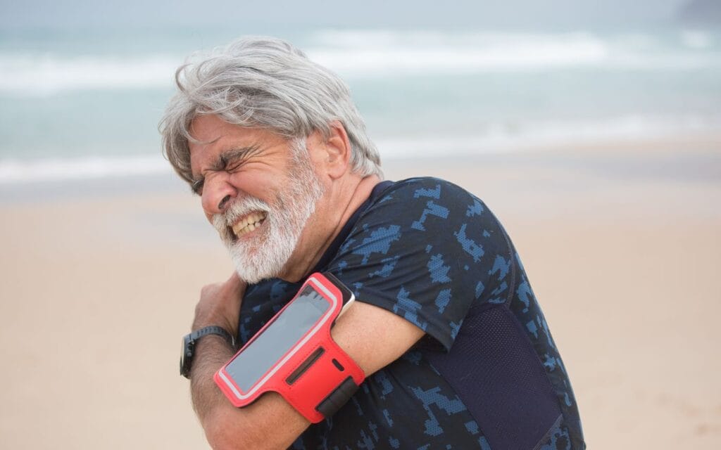 Hombre maduro en la playa sujetándose el hombro por dolor y rigidez (hombro congelado), fisioterapia en Fuengirola